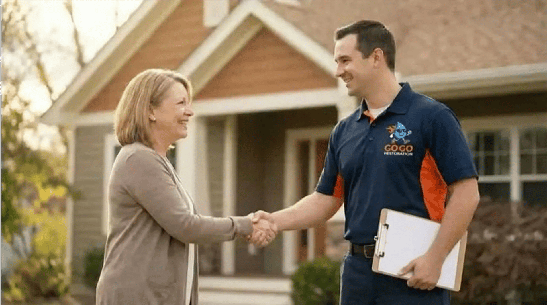 A woman and a man in a GOCO Restoration uniform—former firefighter—shake hands outside a house; the man holds a clipboard, ready to begin property restoration services.